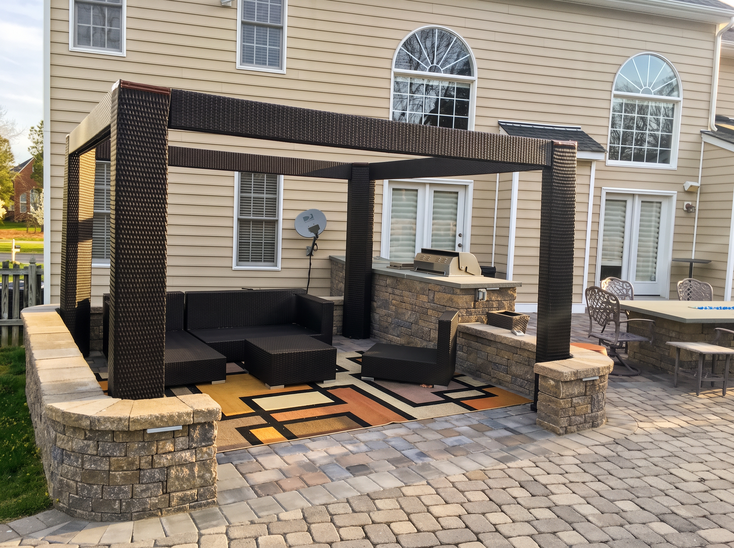 Stone outdoor kitchen and grill under a dark wicker pergola on a paver patio